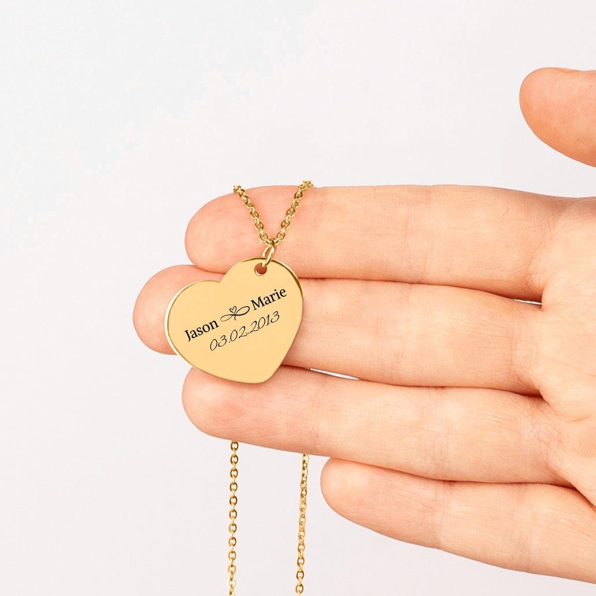 Gold plated heart pendant engraved with two names and a date, held in an open hand
