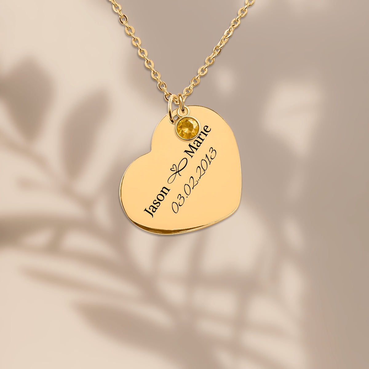Gold plated heart pendant engraved with two names and a date, on a warm sand background