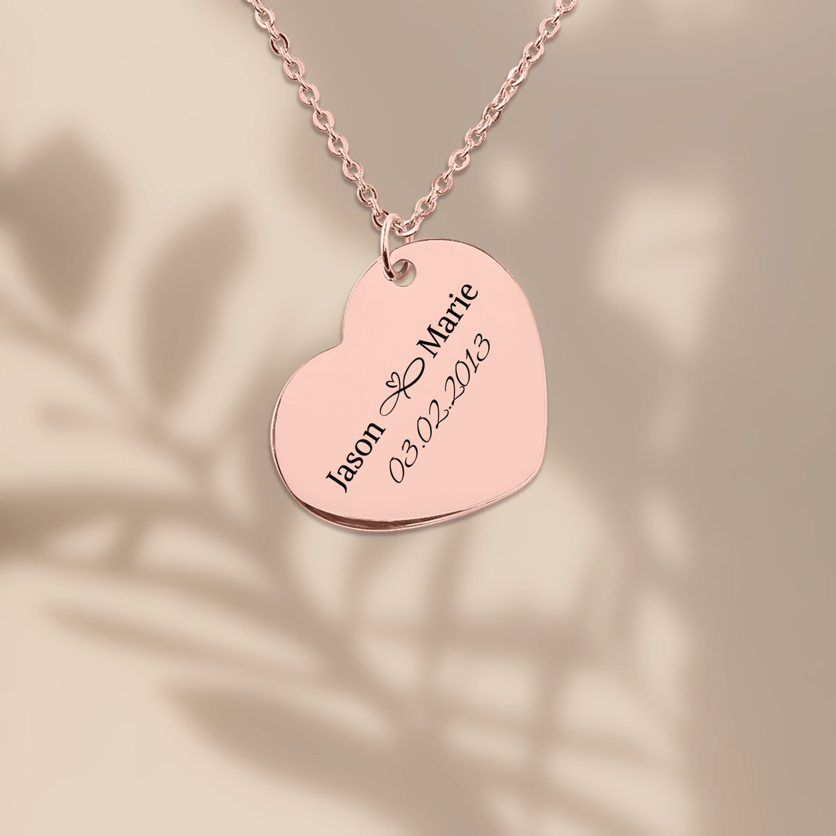 Rose gold plated heart pendant engraved with two names and a date, on a warm sand background